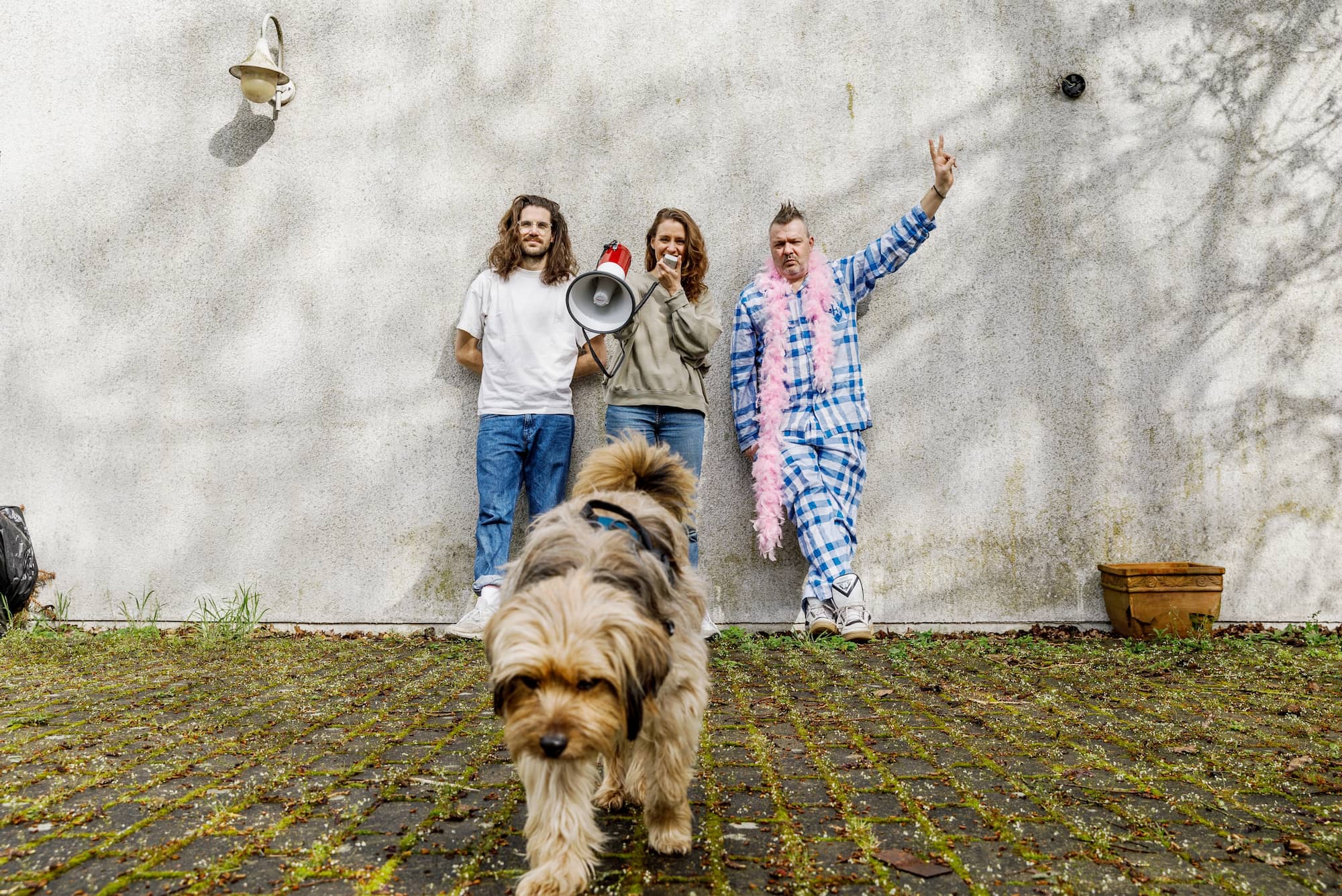 Bandfoto Fortuna Ehrenfeld, die drei Bandmitglieder lehnen an einer weißen Hauswand. Die Person in der Mitte spricht in ein Megafon. Vor der Band steht ein Hund.