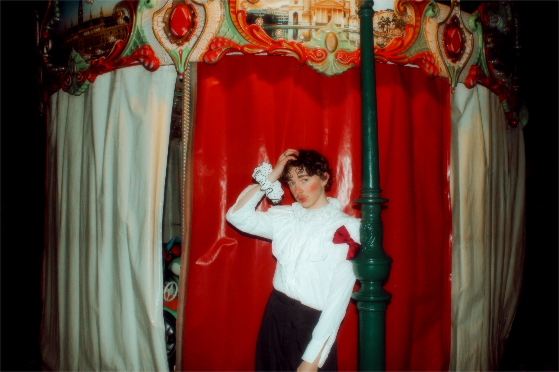 Oskar Haag vor einem bunten Ringelspiel. Er trägt ein weißes Hemd mit Rüschen.